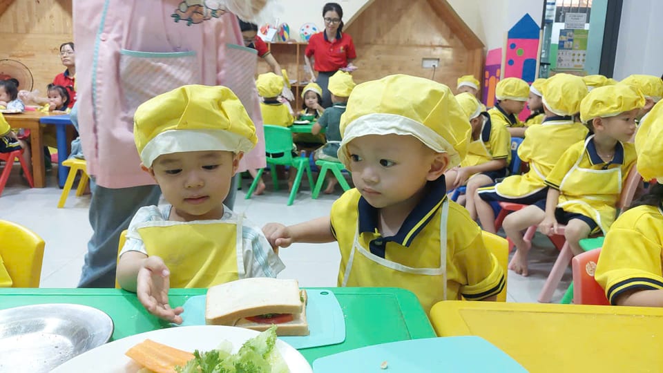 Hai bé đội mũ đầu bếp vàng chăm chú làm bánh sandwich